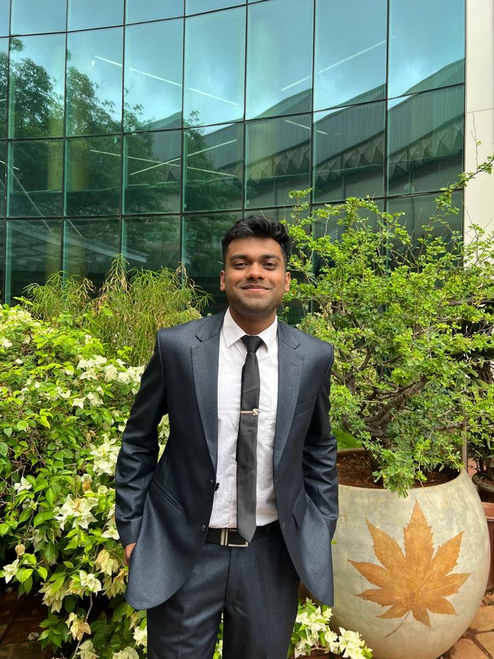 Pravij Gupta in a dark suit, smiling, in front of a glass building and flowering shrubs.